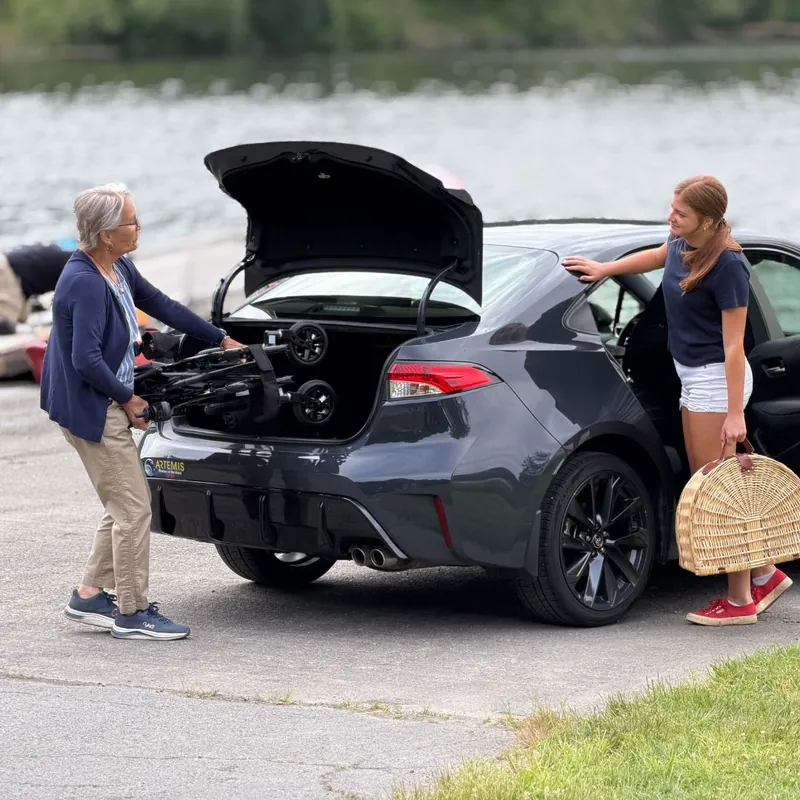 An older woman sliding a folded Golden Cricket LT Carbon power wheelchair into the trunk of a dark gray sedan